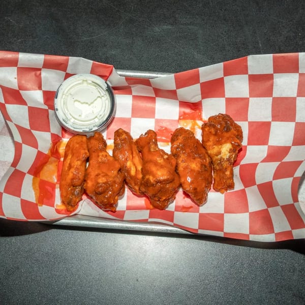Six buffalo wings with ranch dressing in a red and white checkered paper-lined basket.