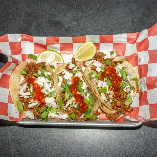 Three street tacos with meat, cabbage, cilantro, red sauce, and lime wedges on checkered paper.