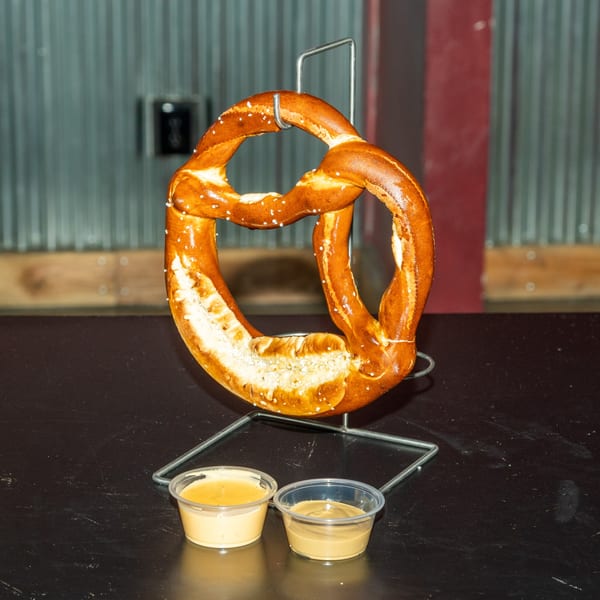 A large, salted pretzel hangs on a metal stand, with two dipping sauces below on a dark table.