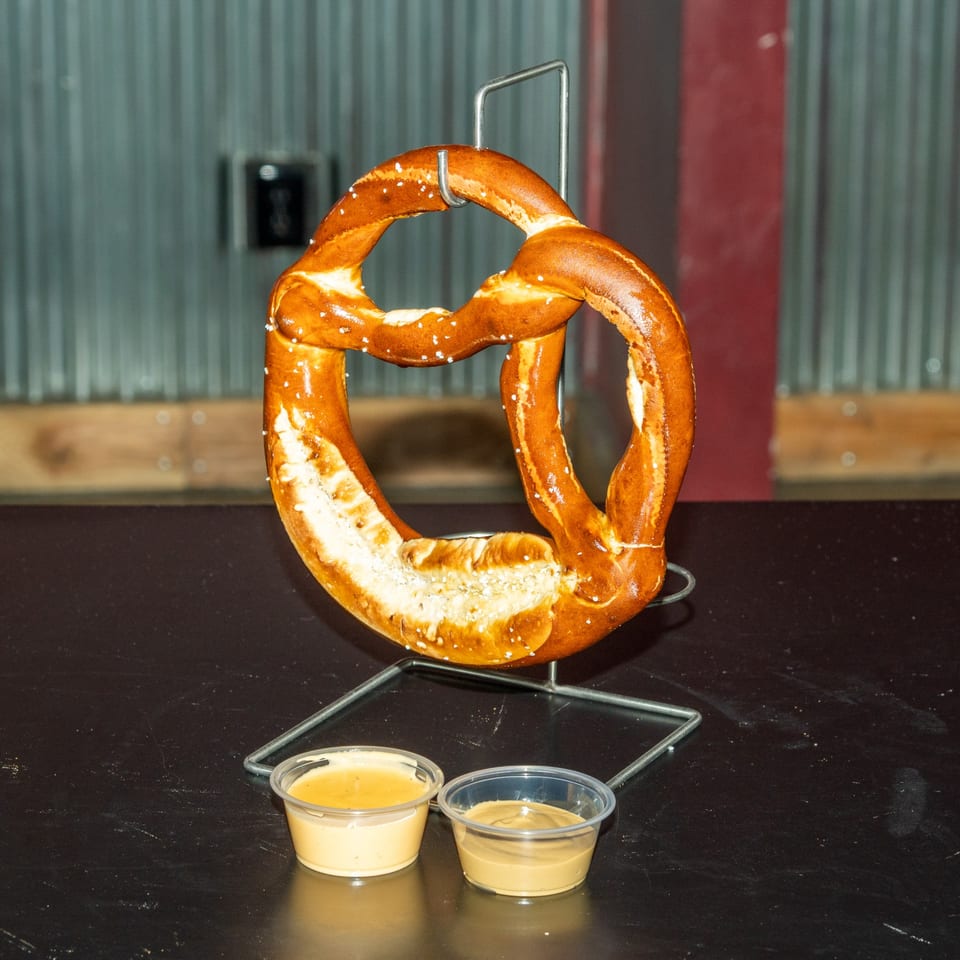 A large, salted pretzel hangs on a metal stand, with two dipping sauces below on a dark table.