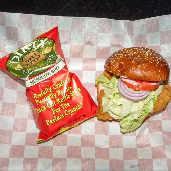 A fried chicken sandwich with lettuce, tomato, and onion, next to a bag of Dirty Mesquite BBQ chips.