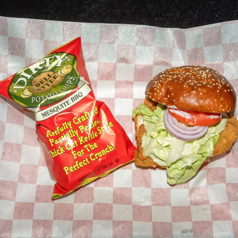 A fried chicken sandwich with lettuce, tomato, and onion, next to a bag of Dirty Mesquite BBQ chips.