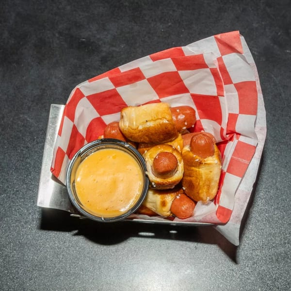 Pigs in a blanket with pretzel dough and a side of cheese dipping sauce in a metal basket.