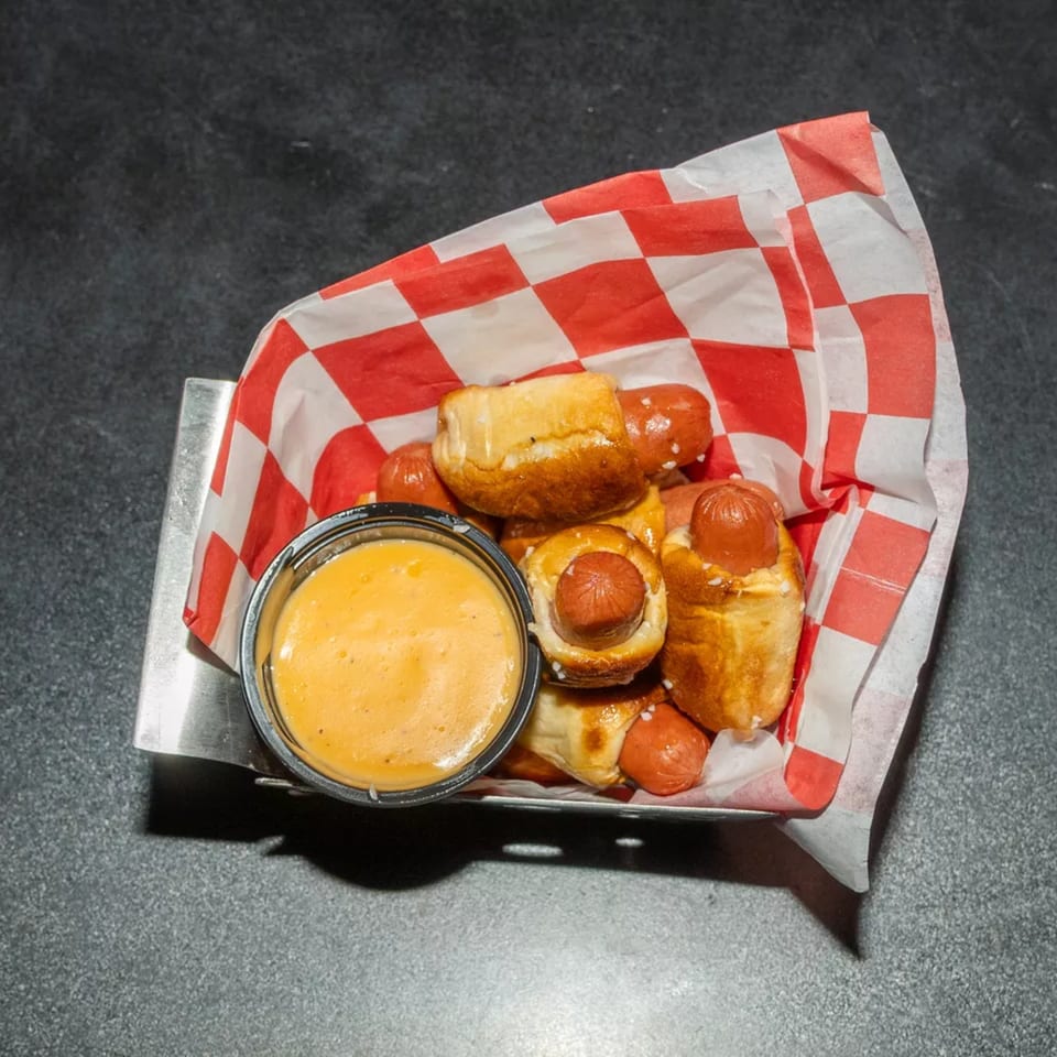 Pigs in a blanket with pretzel dough and a side of cheese dipping sauce in a metal basket.